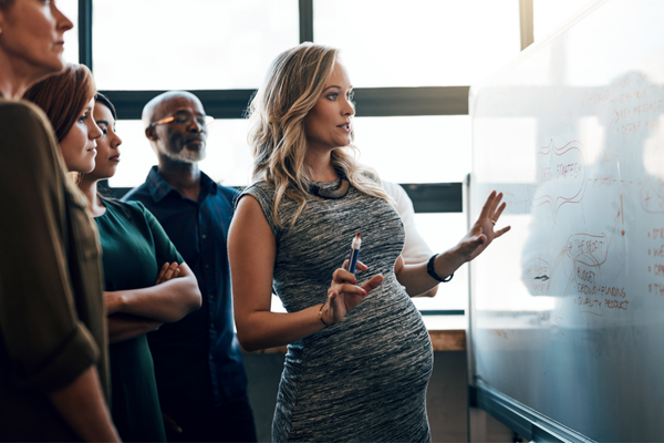 A pregnant woman leads a workplace discussion in front of a whiteboard.